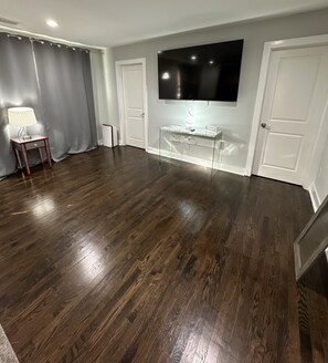 Ein geräumiges Zimmer mit einem großen Flachbildfernseher an der Wand, einem kleinen Tisch mit Lampe und einer Tür rechts.