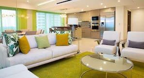A modern living room with a sofa, armchairs, a glass coffee table, and a kitchen area with appliances.