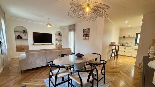 Un soggiorno moderno con divano, tavolo da pranzo e sedie, una TV fissata al muro e una zona cucina sullo sfondo.