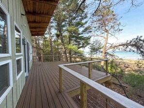 A wooden deck with a railing, overlooking a forested area and a body of water. - Burns No.8 - Cottage on the Bay (Kewadin)