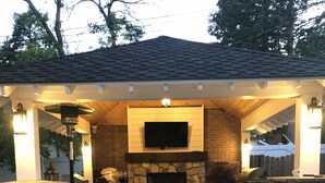 Un patio al aire libre con una chimenea de piedra, un televisor de pantalla plana montado encima y una mesa de comedor con sillas.