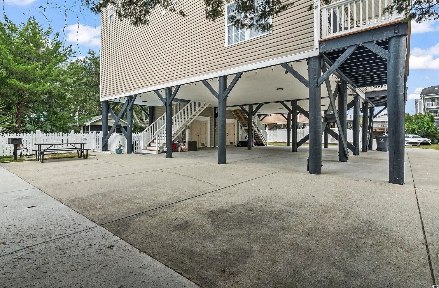 Un edificio con un espacio exterior cubierto sostenido por vigas metálicas, una escalera y una cerca de piquetes blancos.