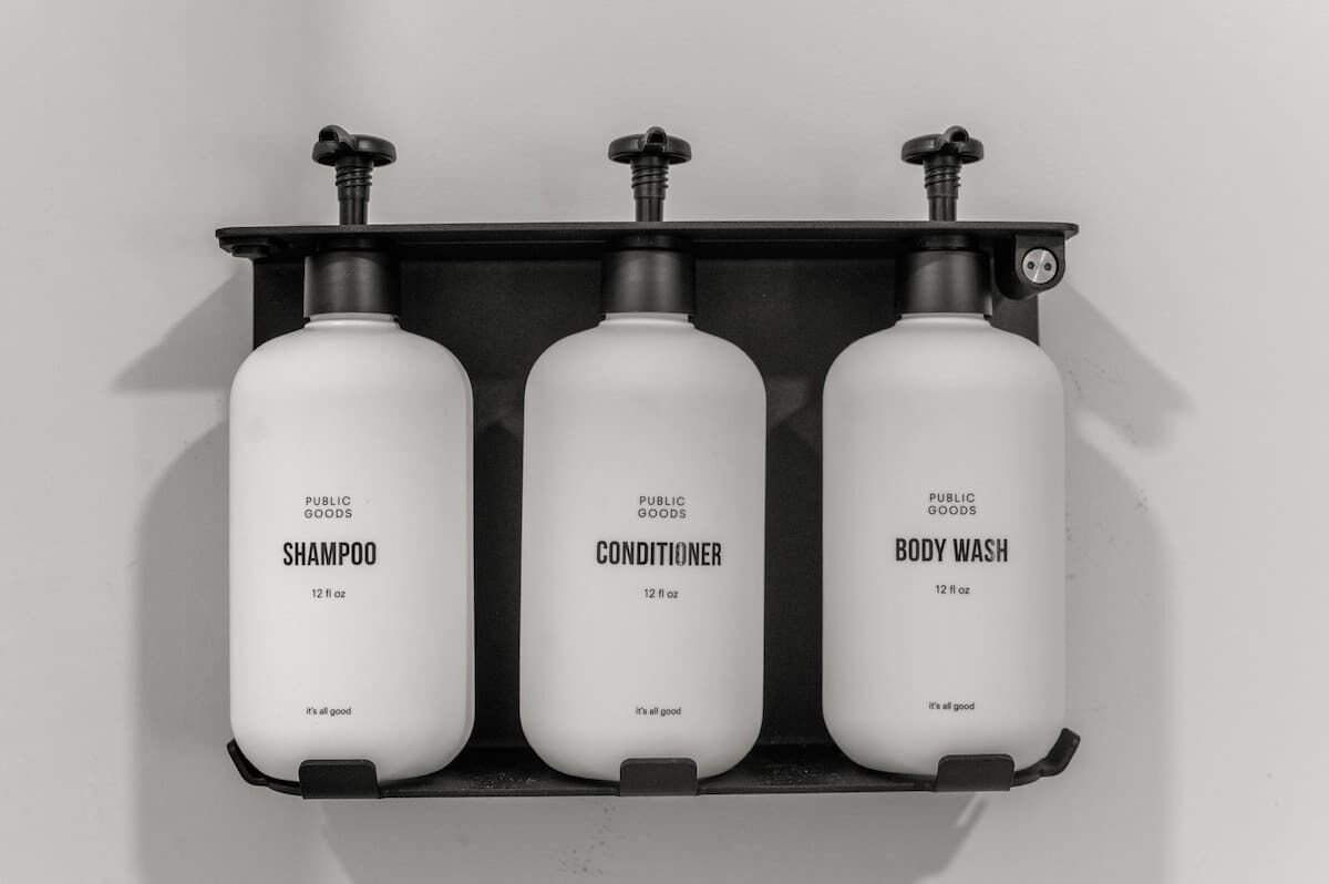 Tre hvide flasker mærket 'Public Goods' står på en sort hylde. Hver flaske er markeret med '12 fl oz' og teksten 'It's all good'.