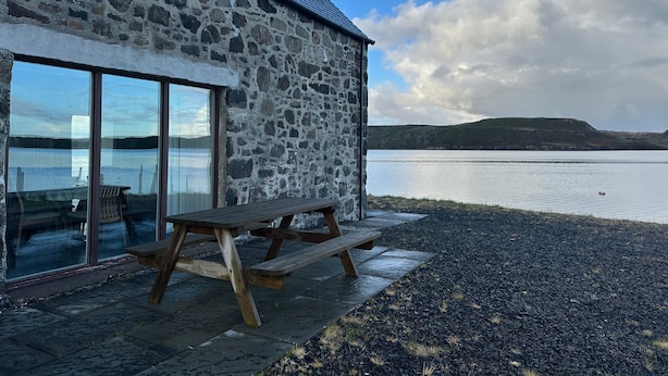 A stone building with a wooden picnic table and a glass door leading to a seating area. - A secluded Skye boathouse where rugged beauty meets refined coastal living. (Skye)