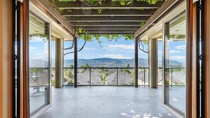 Un balcón cubierto con vistas a montañas distantes, una pérgola con hiedra y una puerta de cristal.
