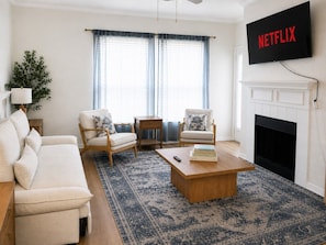 A living room with a sofa, two armchairs, a coffee table, a fireplace, and a television displaying 'NETFLIX'. - Designer Dream 3BR for Families & LongStays + Pool (Plano)