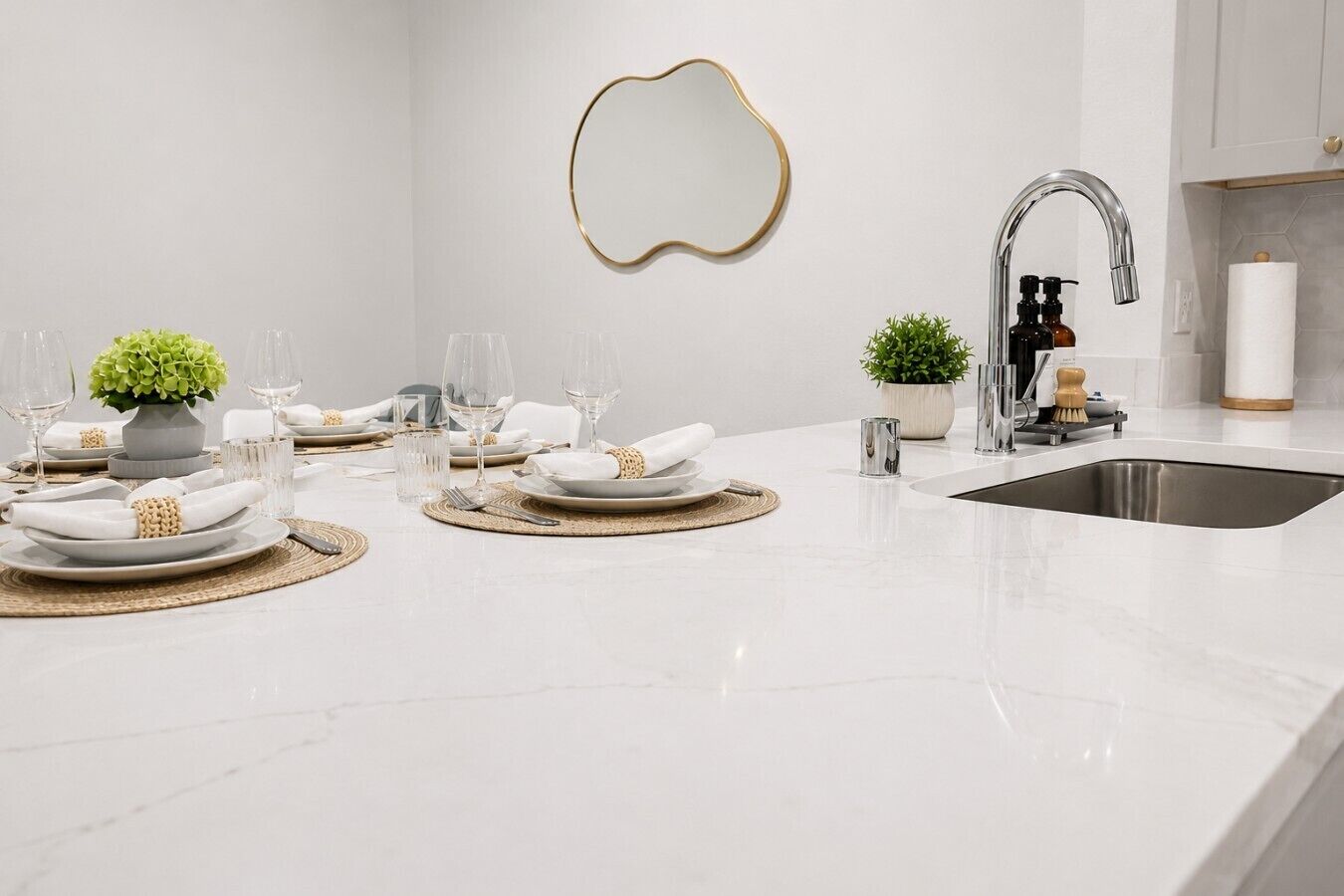A modern kitchen with a white countertop, a stainless steel sink, and a decorative mirror.