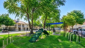 Un parque infantil con tobogán y columpios, rodeado de árboles y una valla.