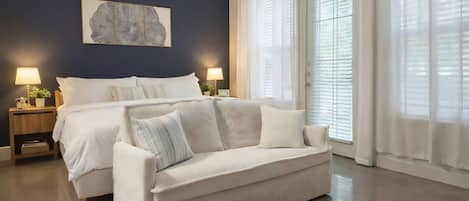 A bedroom with a bed, a white sofa, two bedside tables with lamps, a large window with blinds, and a wall-mounted artwork.