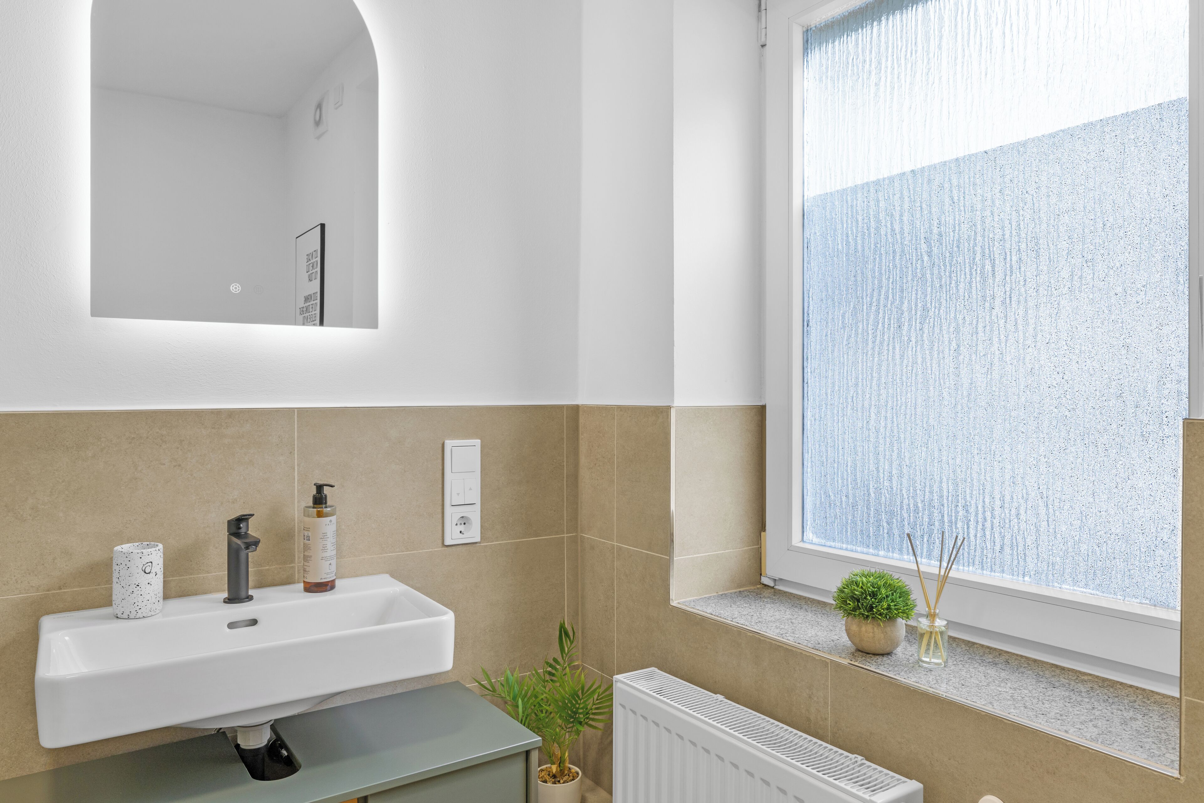 Een badkamer met een witte wastafel, een spiegel, een potplant en een raam met uitzicht.
