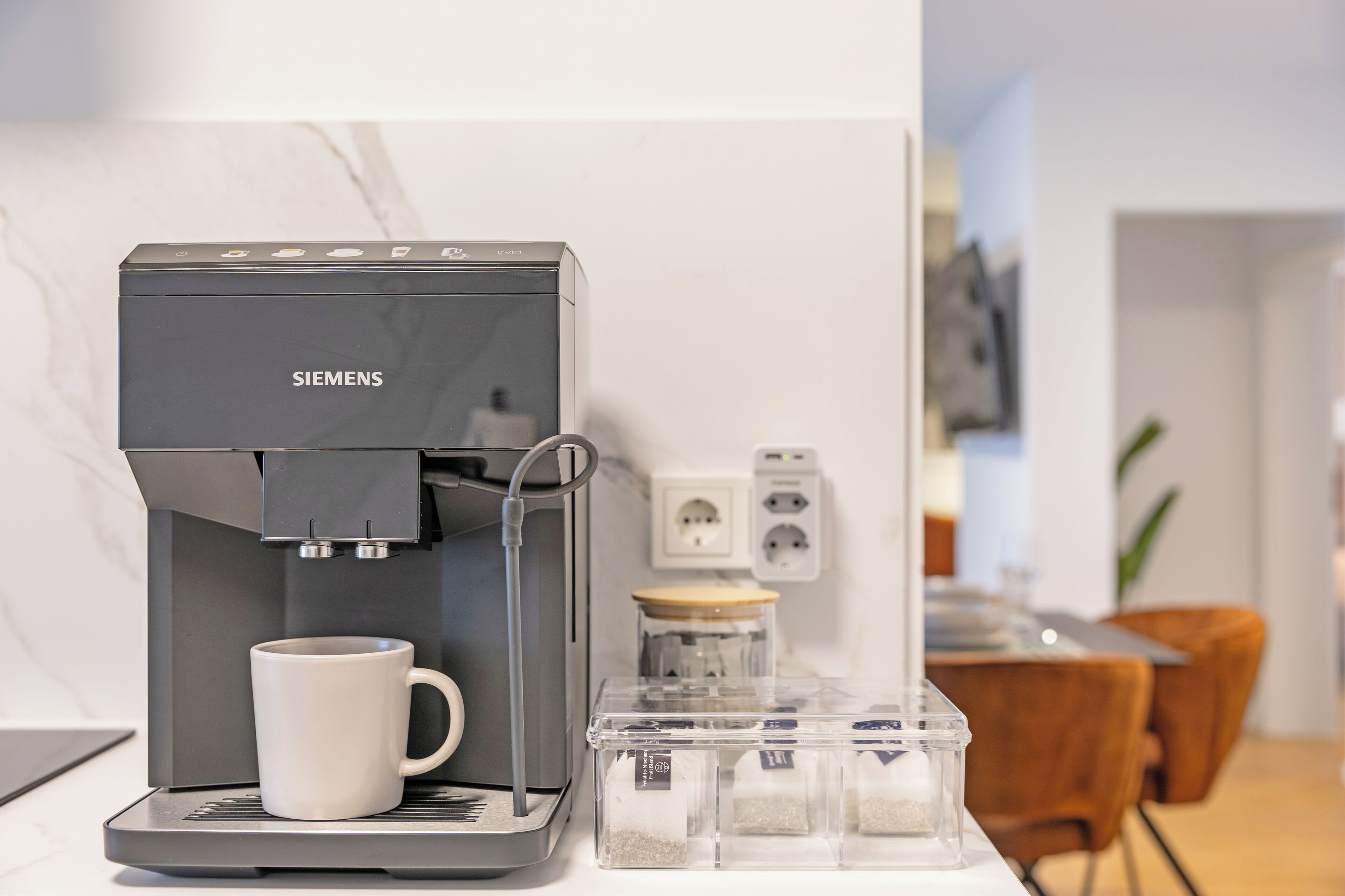 A Siemens coffee machine with a white mug, a clear container with coffee pods, and a power outlet in a modern kitchen setting.