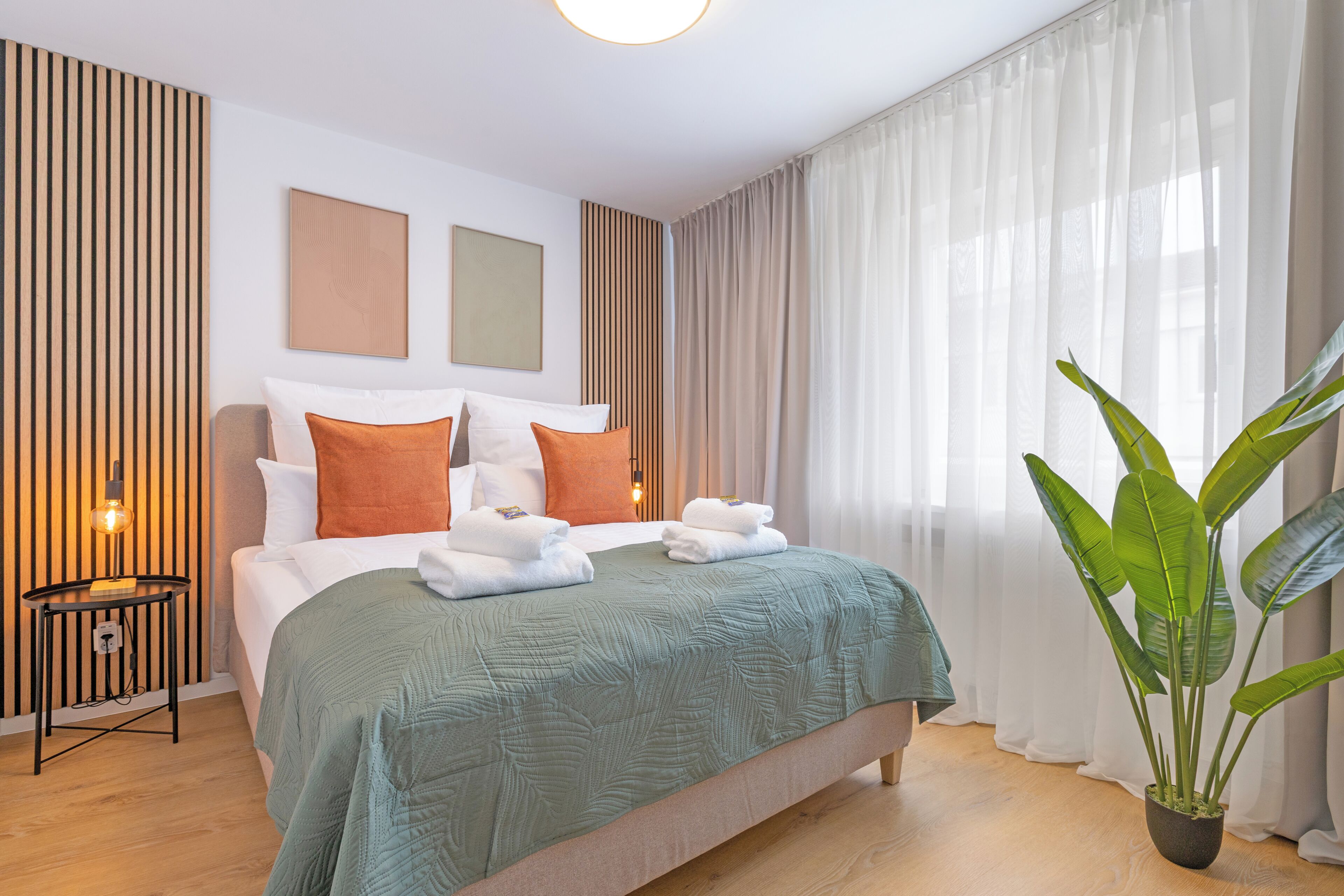 A hotel room with a bed, a nightstand, a potted plant, and a window with curtains.