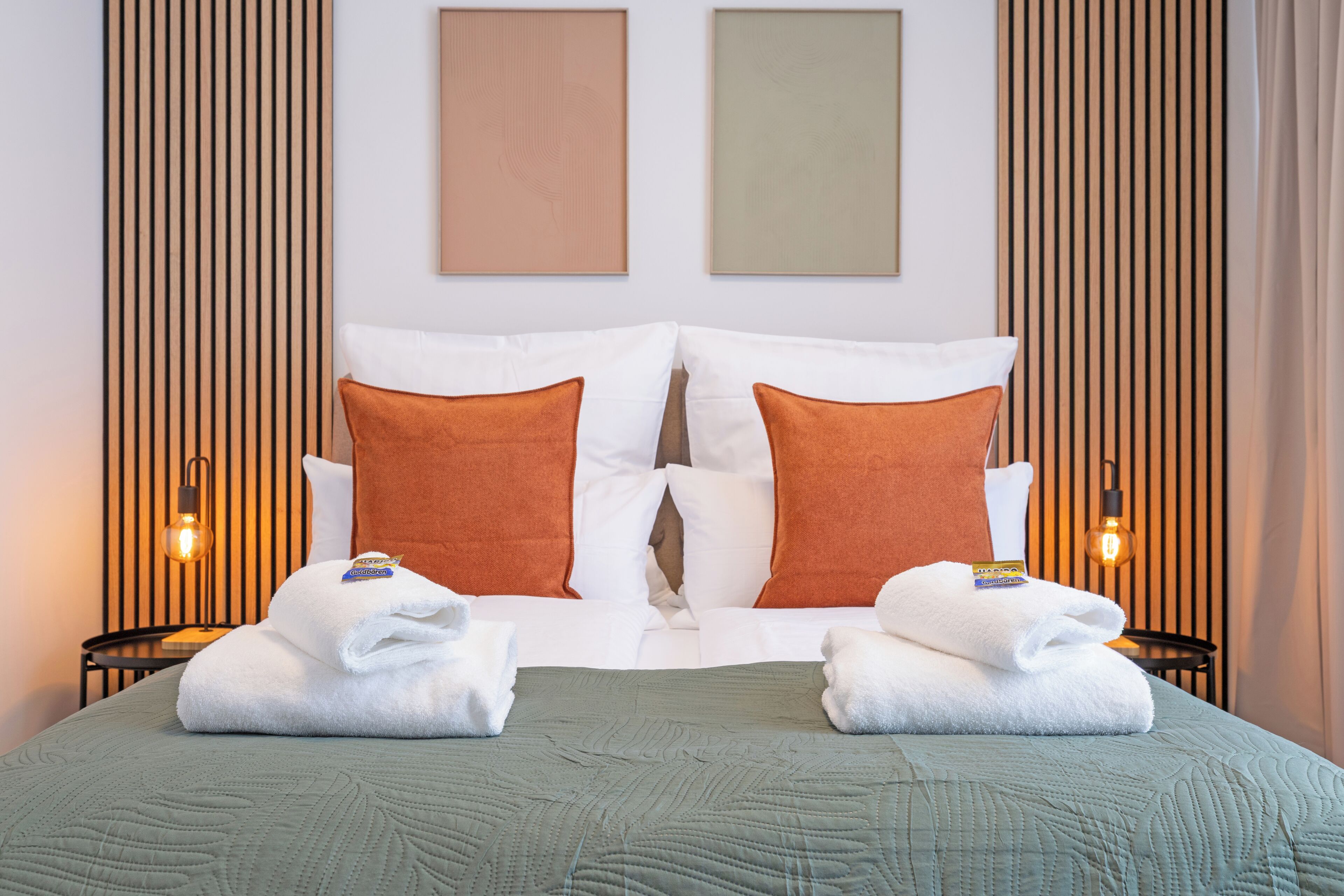 A neatly made bed with white and orange pillows, a green quilt, and two towels on the nightstands.