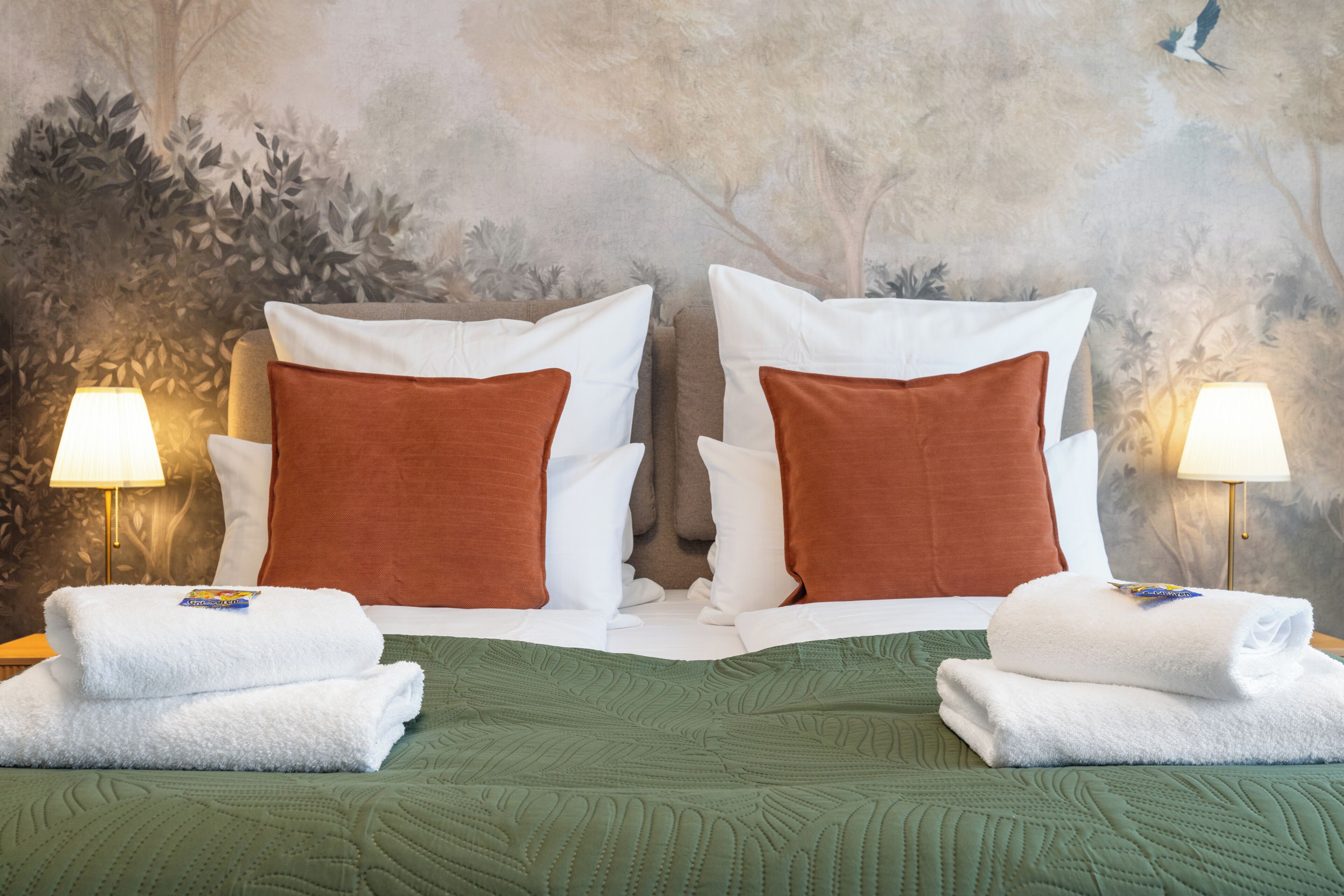 Cama doble con ropa de cama verde, dos almohadas blancas y dos almohadas marrones. Hay dos lámparas de noche y dos toallas blancas dobladas sobre la cama.