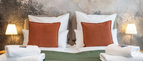 Cama doble con ropa de cama verde, dos almohadas blancas y dos almohadas marrones. Hay dos lámparas de noche y dos toallas blancas dobladas sobre la cama.
