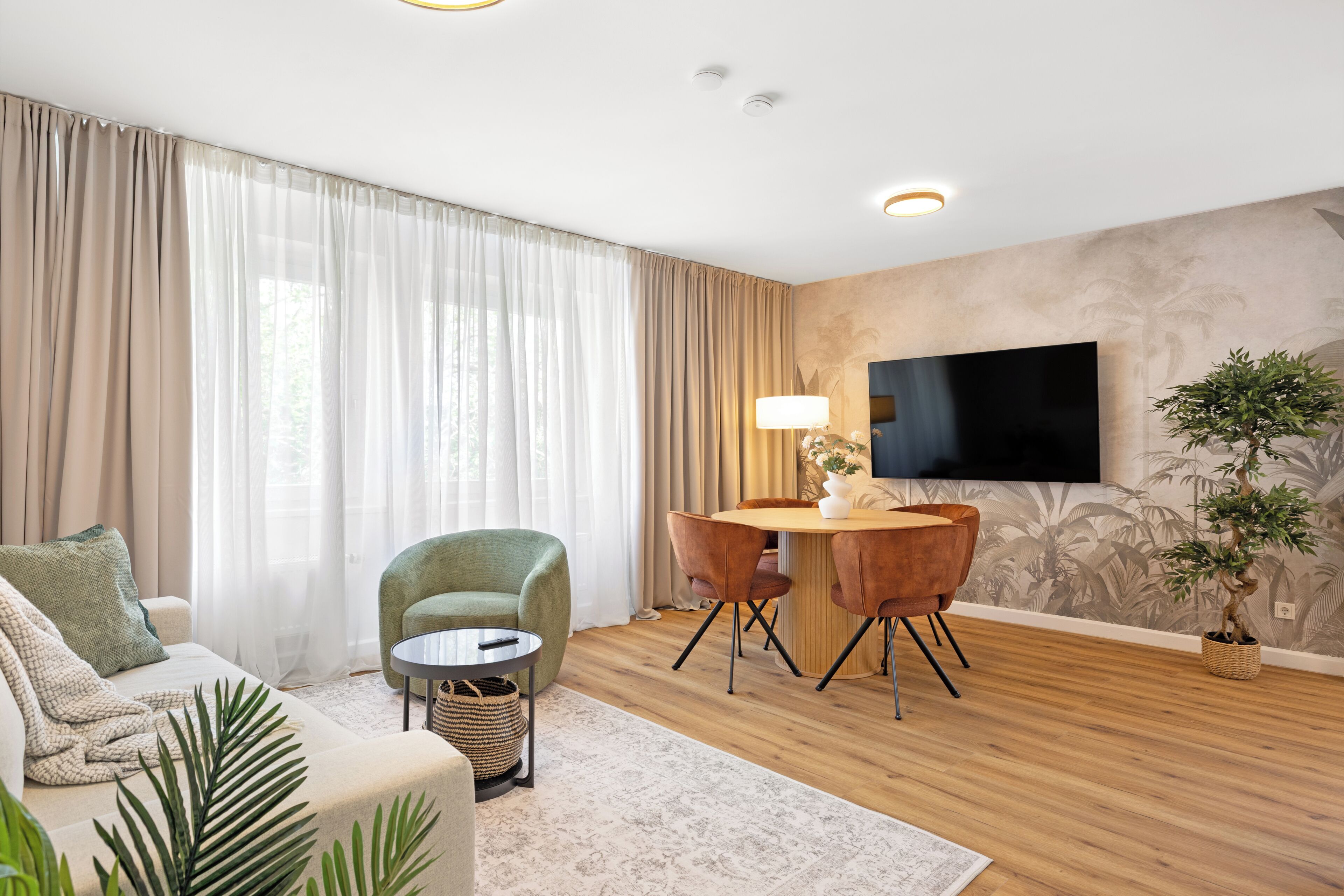 A living room with a sofa, a green armchair, a round table with chairs, a TV, and a plant.