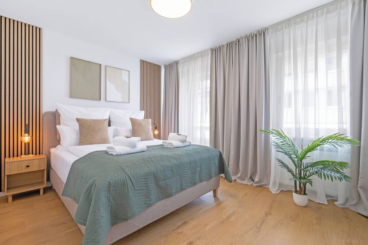 A modern hotel room with a bed, bedside tables, a potted plant, and a large window with curtains.