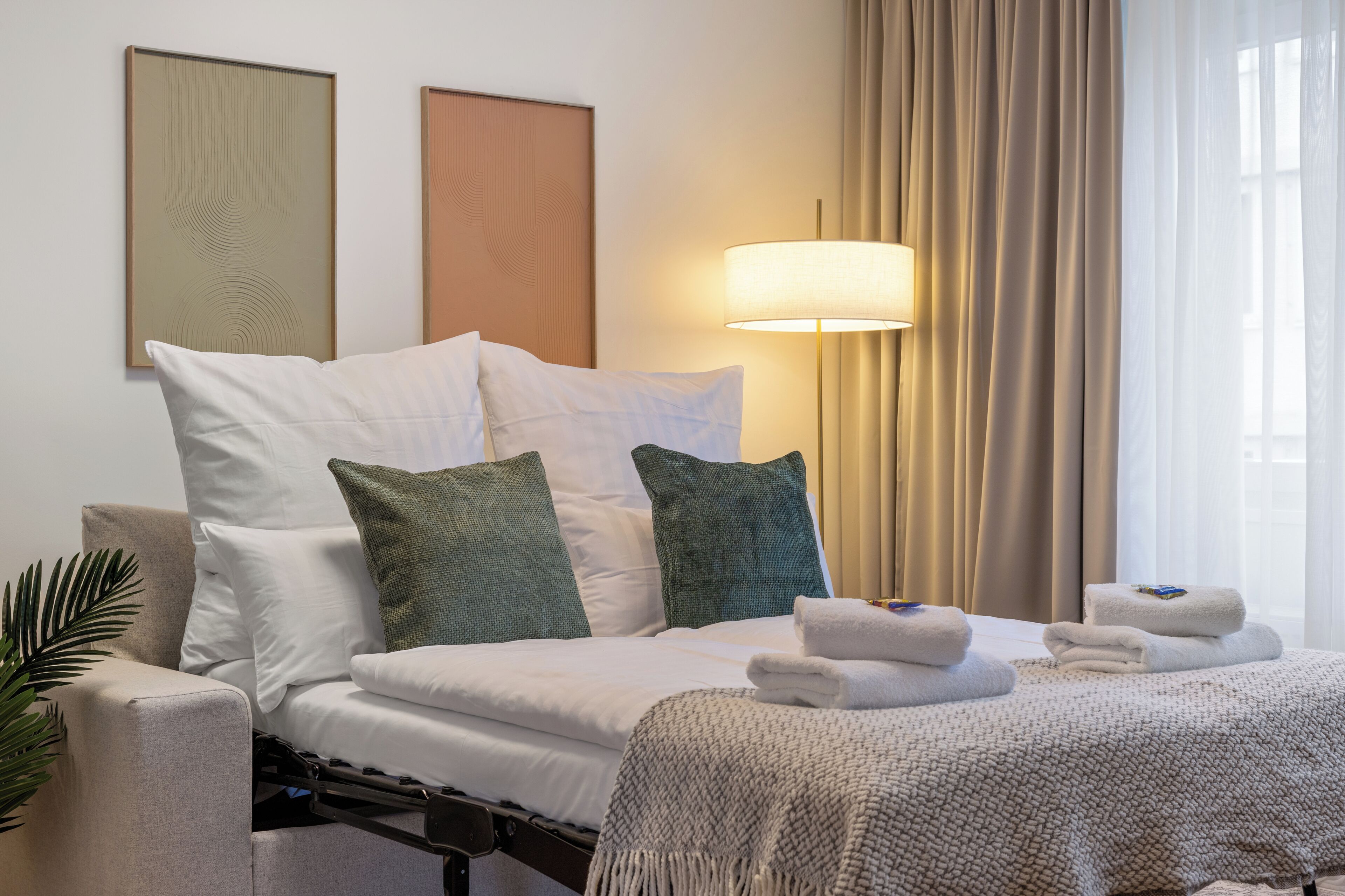 A hotel room with a bed, pillows, a lamp, curtains, and a plant.