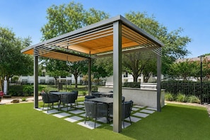 Eine überdachte Terrasse im Freien mit Tischen und Stühlen, umgeben von einem gepflegten Rasen und Bäumen.