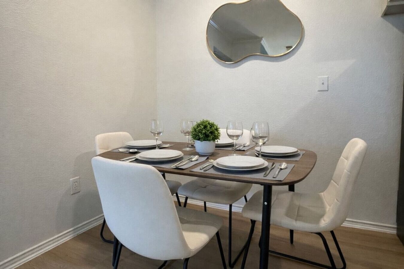 A dining area with a table set for four, white chairs, a potted plant, and a wall-mounted mirror.