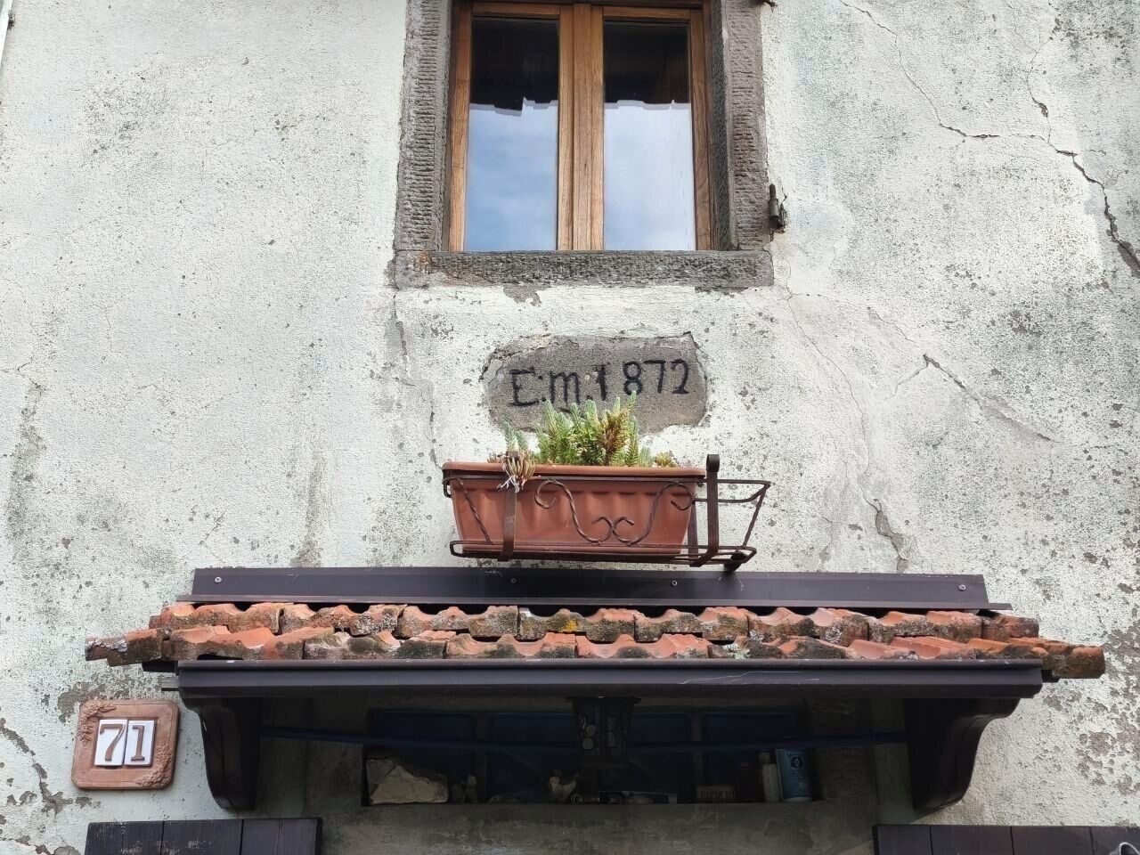 Ein Gebäude mit einem Fenster, einer Tafel mit der Inschrift 'E.M. 1872', einem Backsteinvorsprung und einem Blumentopf.