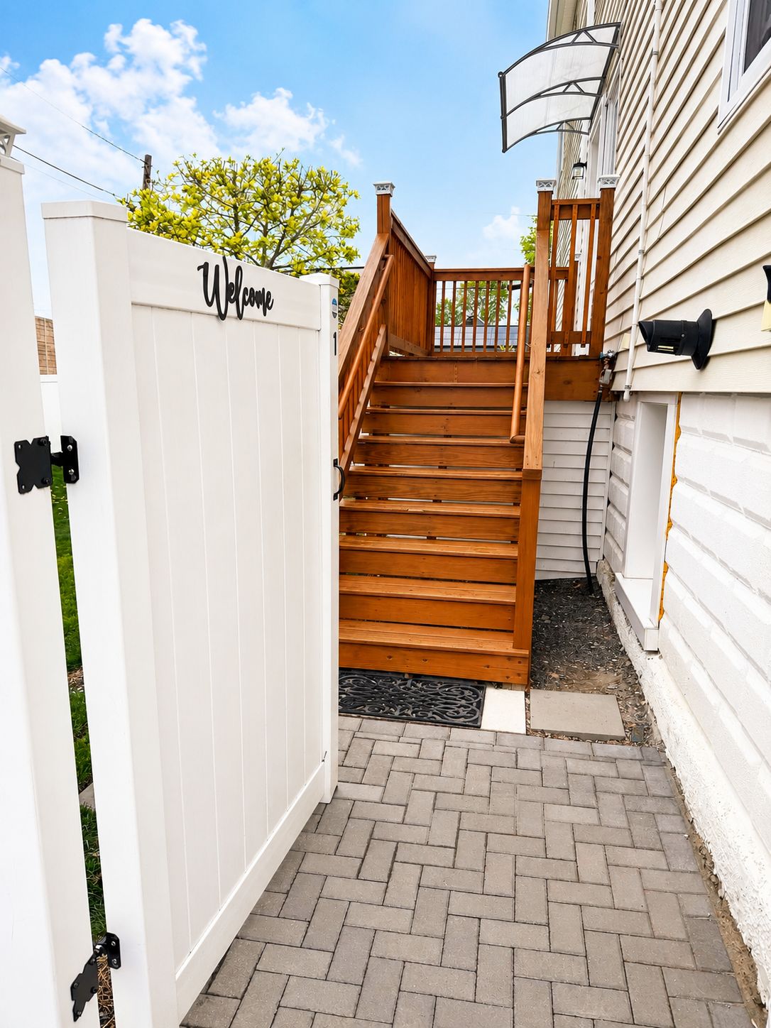 Una escalera de madera que lleva a un portón blanco con un letrero que dice 'Bienvenido', un sendero de ladrillos y una casa con revestimiento.
