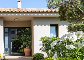 Ein Haus mit Ziegeldach, einer Tür mit Glasfeldern und einem Fenster. Im Vordergrund befinden sich ein Blumentopf und weiße Blumen.