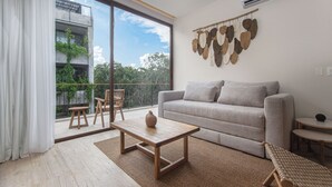 En moderne stue med sofa, sofabord og lænestole, et stort vindue med udsigt til grøn vegetation, samt en vægmonteret klimaanlæg.