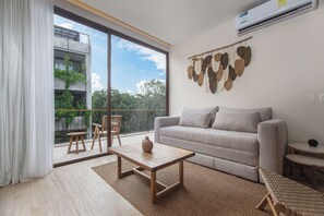 Ein modernes Wohnzimmer mit einer Couch, einem Couchtisch und Sesseln, einem großen Fenster mit Blick ins Grüne und einer Wand-Klimaanlage.