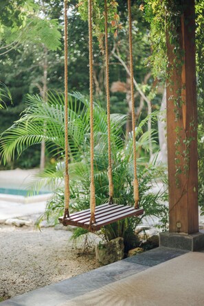 Eine Holzliege, an Seilen aufgehängt, umgeben von üppiger Vegetation und einem Pool.