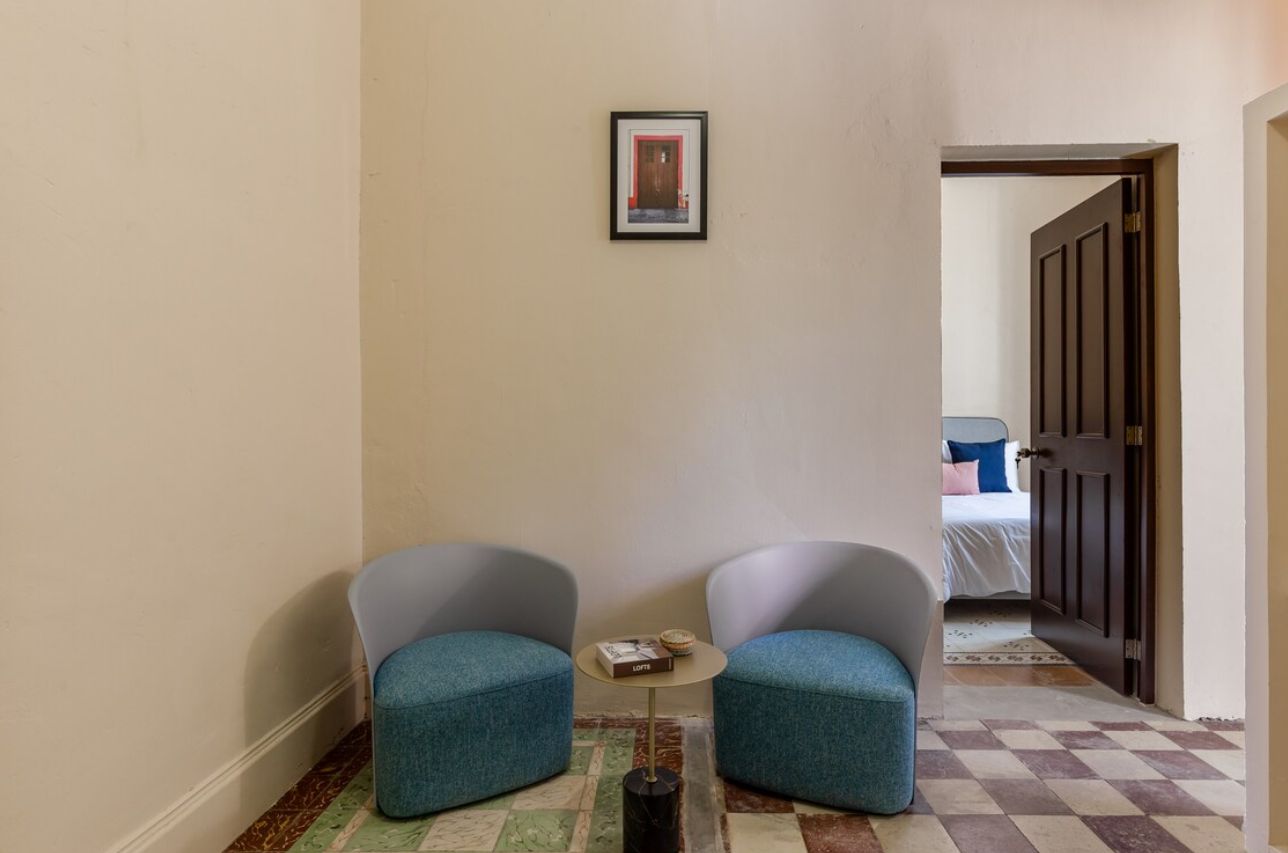 Un couloir avec deux fauteuils rembourrés, une petite table sur laquelle se trouve une boîte, et une porte donnant sur une chambre.