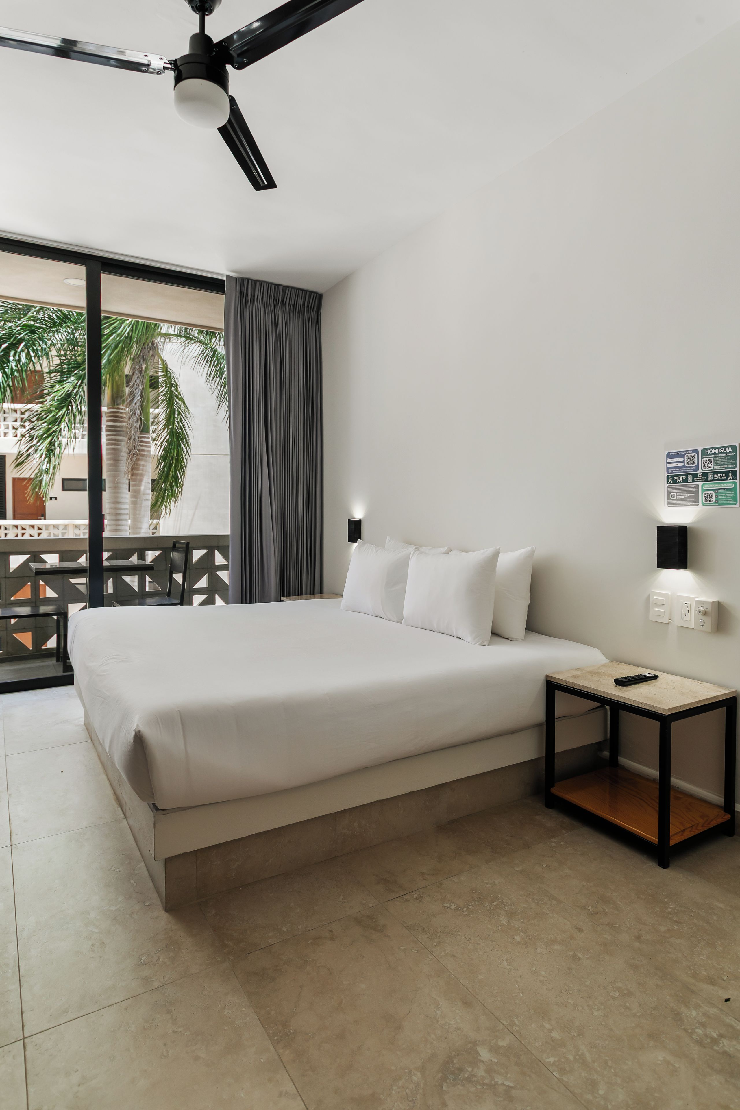 A hotel room with a bed, a nightstand, a remote control, a wall-mounted light, and a view of palm trees through the window.