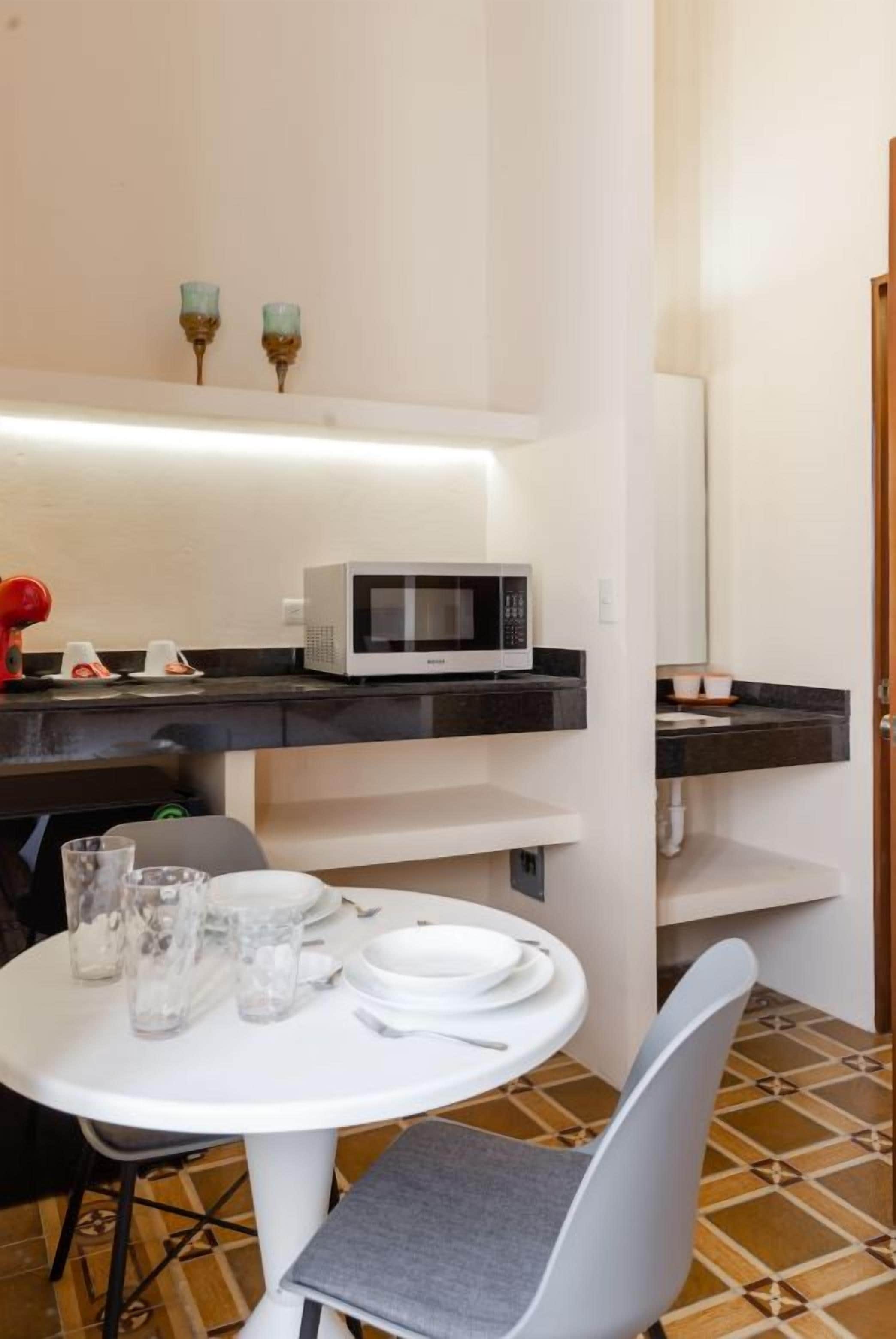 Een kleine kitchenette met een magnetron, een witte tafel met stoelen en een betegelde vloer.