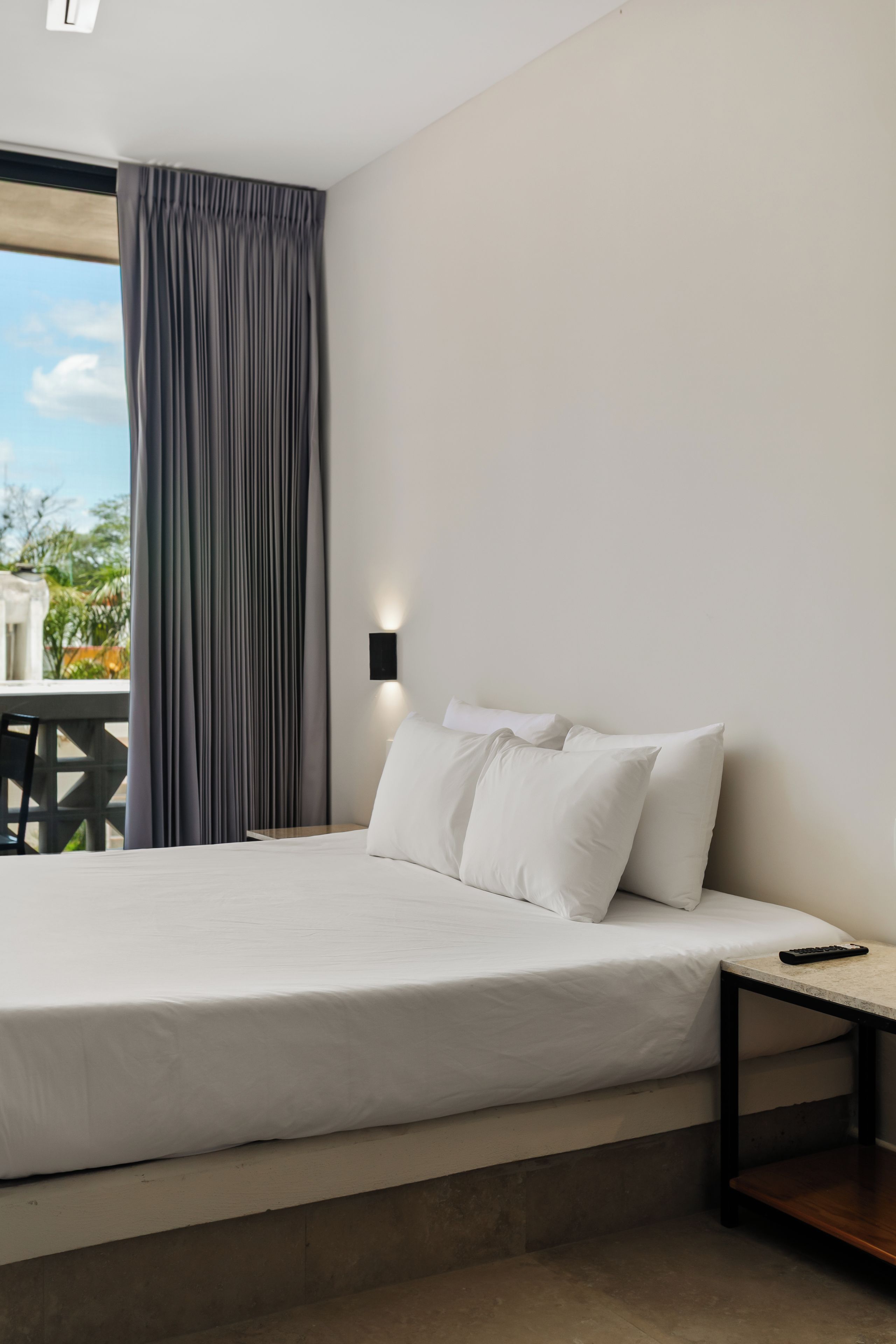 A bedroom with a bed, a nightstand, a wall-mounted light, and a view of the outdoors.