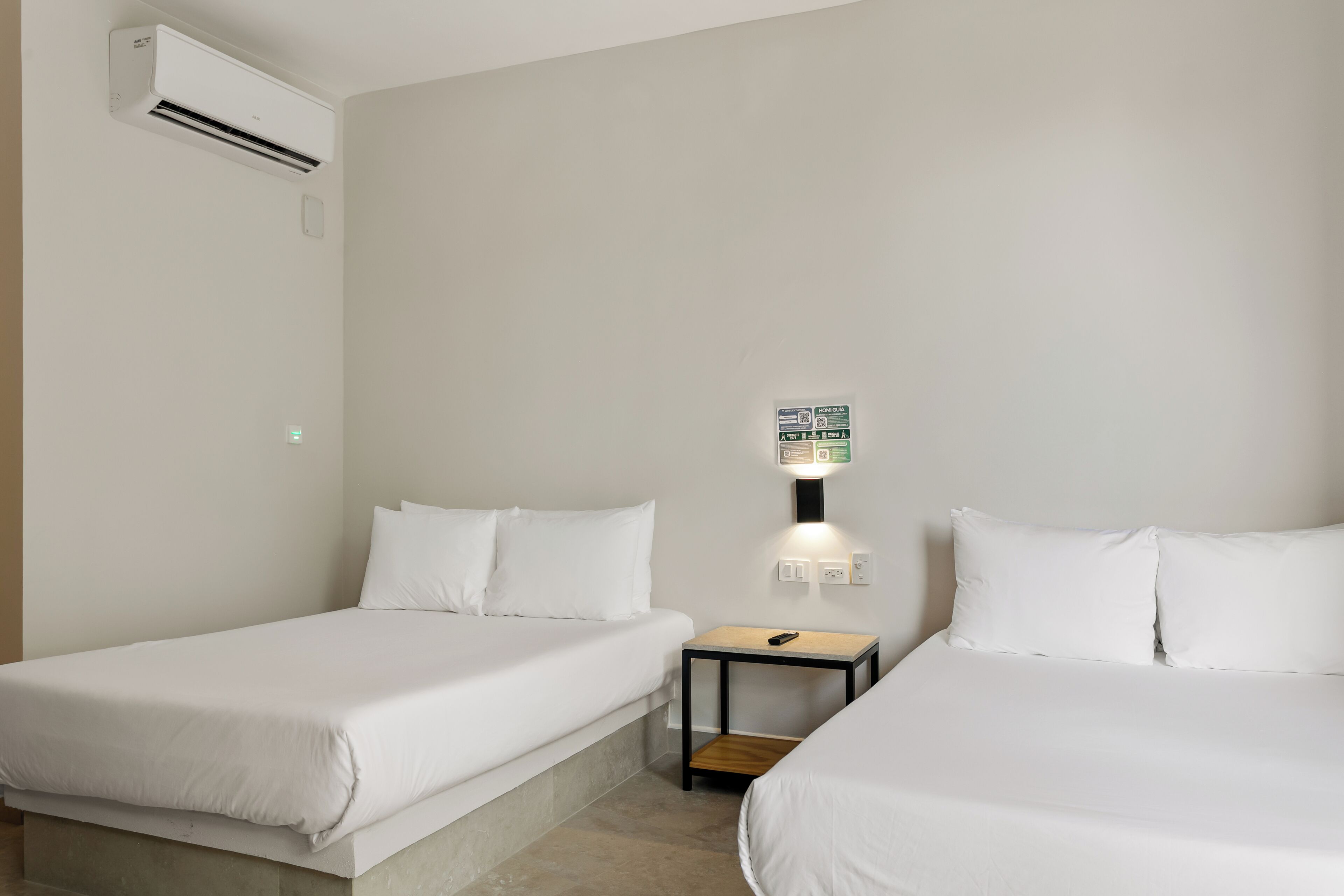 A hotel room with two single beds, a bedside table with a lamp, an air conditioning unit, and a wall-mounted control panel.