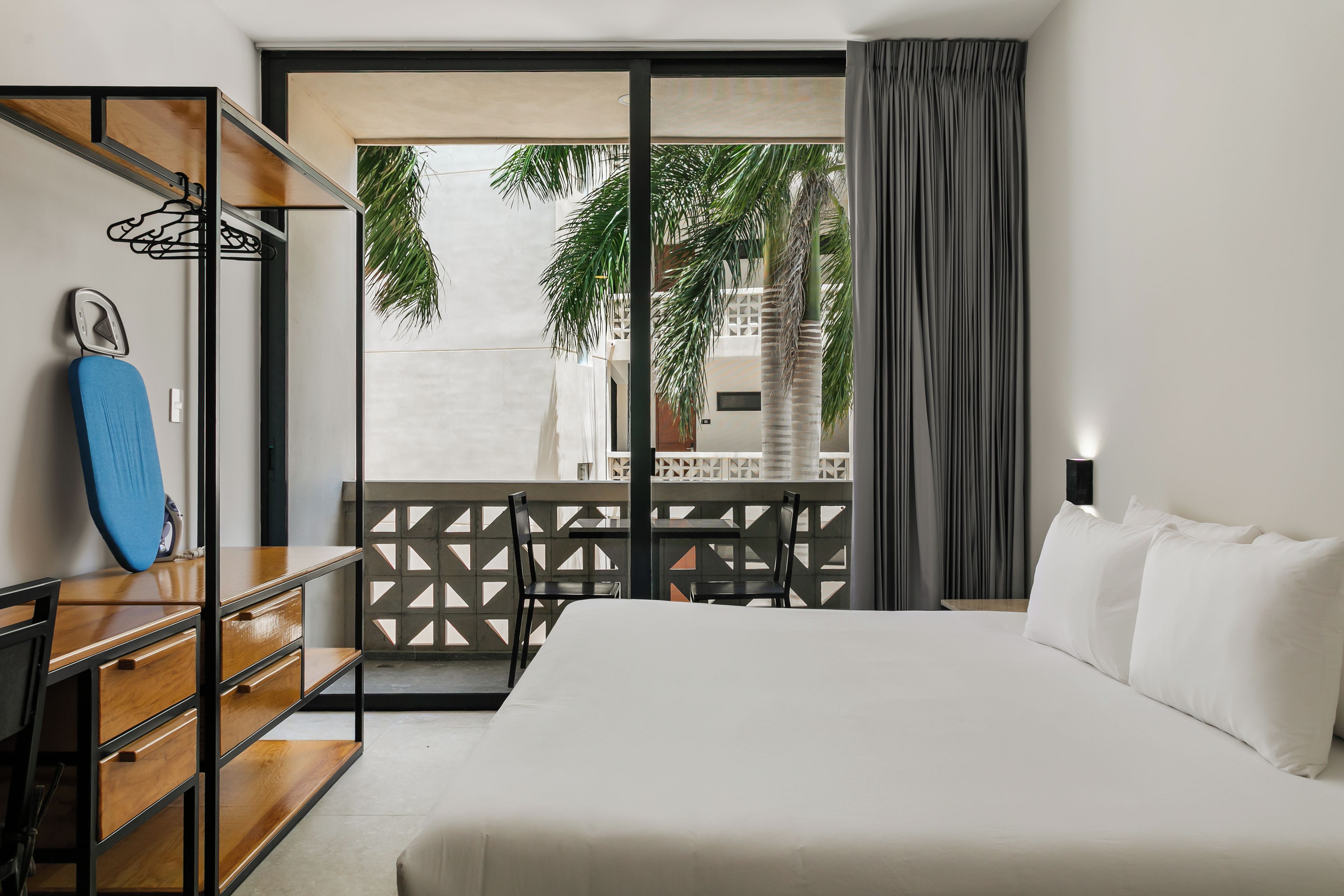 A hotel room with a bed, a desk, a chair, a wardrobe, and a view of palm trees through a balcony.