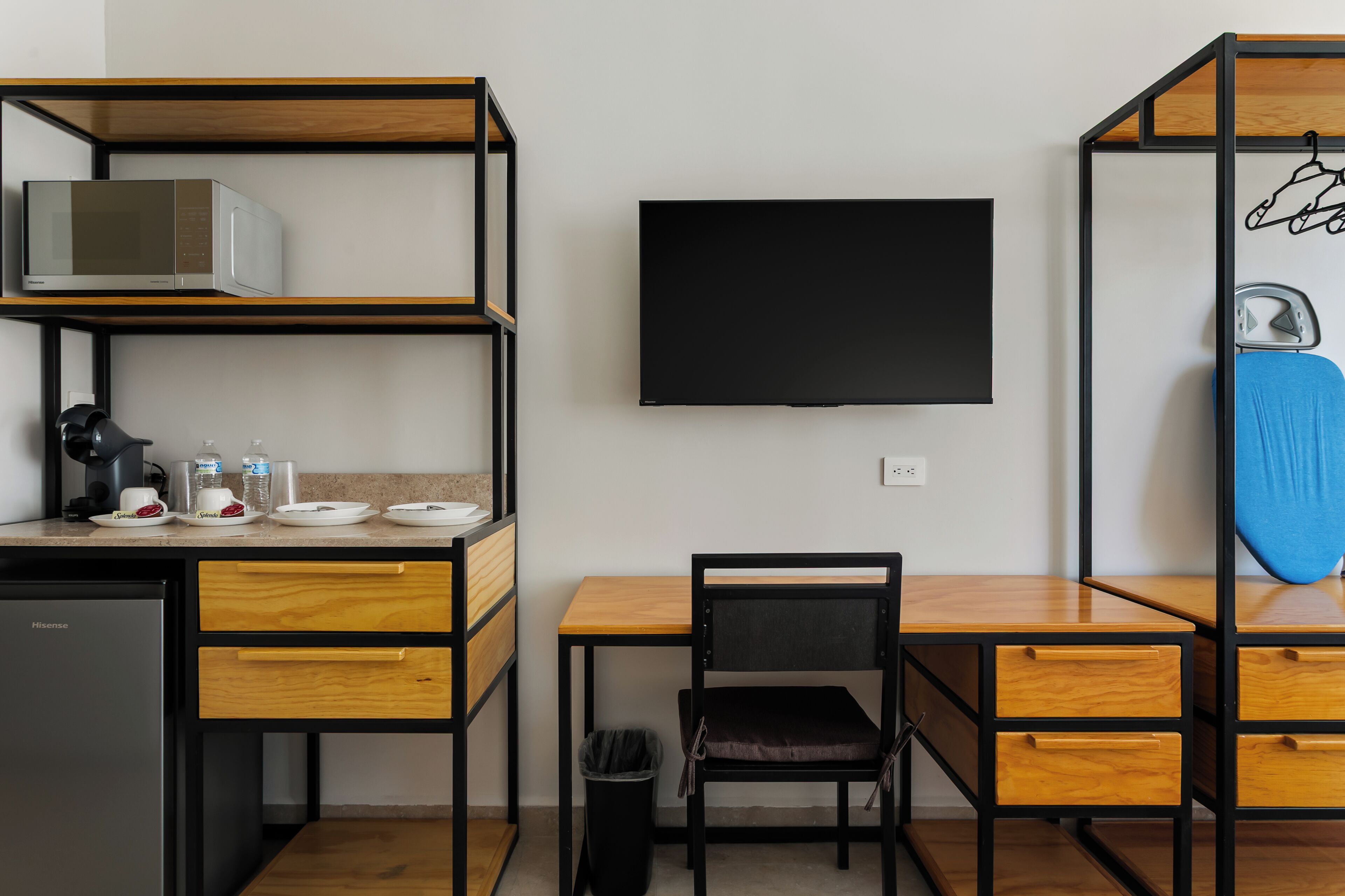 Une pièce compacte comprenant un coin cuisine, un téléviseur à écran plat, un bureau en bois, une chaise et une petite poubelle.
