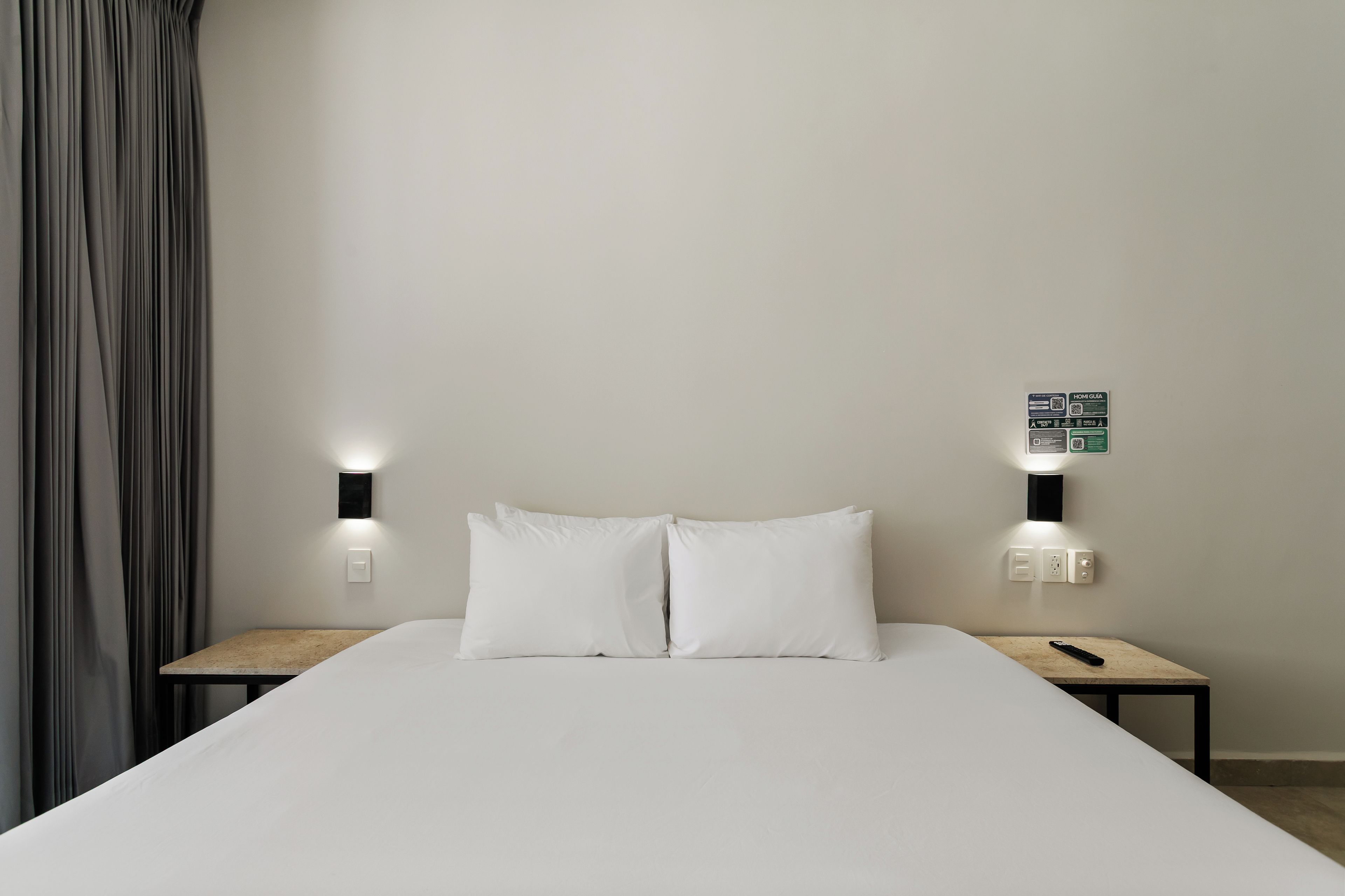 Una habitación de hotel con una cama, dos mesitas de noche, un control remoto y un interruptor de luz en la pared.