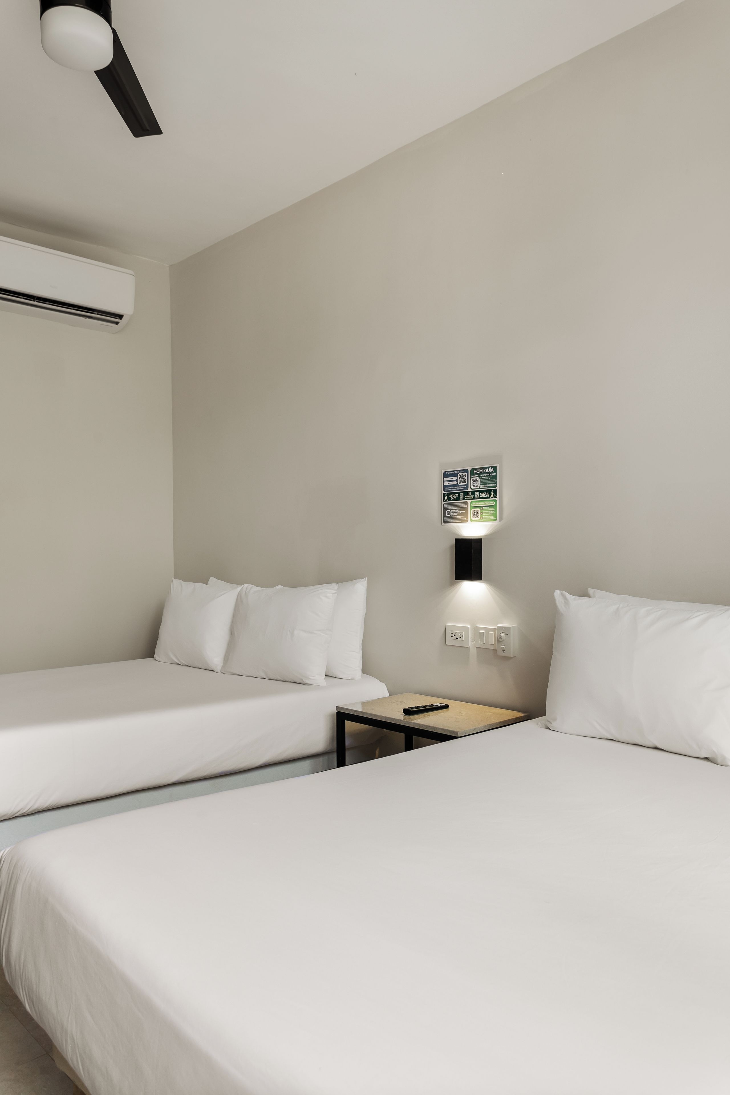 Ein Hotelzimmer mit zwei Betten, einer wandmontierten Klimaanlage, einem Nachttisch mit einer Fernbedienung und einer Wandleuchte.