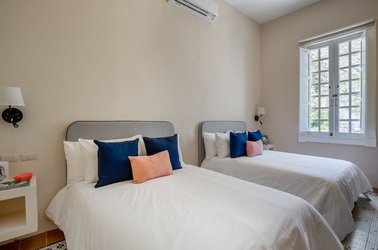 A hotel room with two beds, a window, and an air conditioning unit.