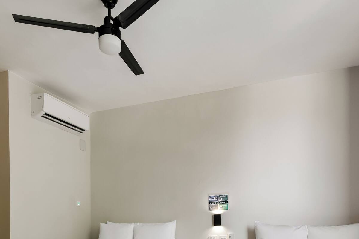 A hotel room with two beds, a ceiling fan, an air conditioning unit, a bedside table with a lamp, and a wall-mounted sign.