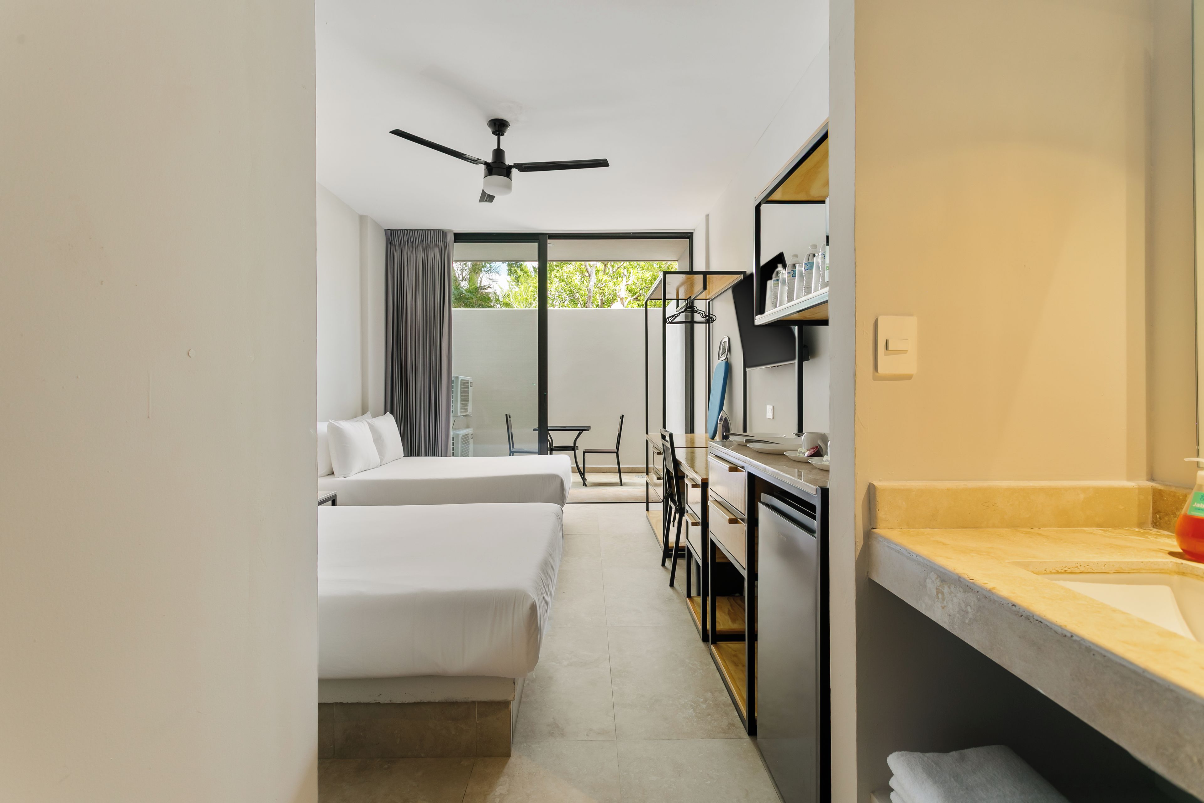 Une chambre d’hôtel avec deux lits, un bureau, une chaise, une salle de bain avec un lavabo et un ventilateur de plafond.