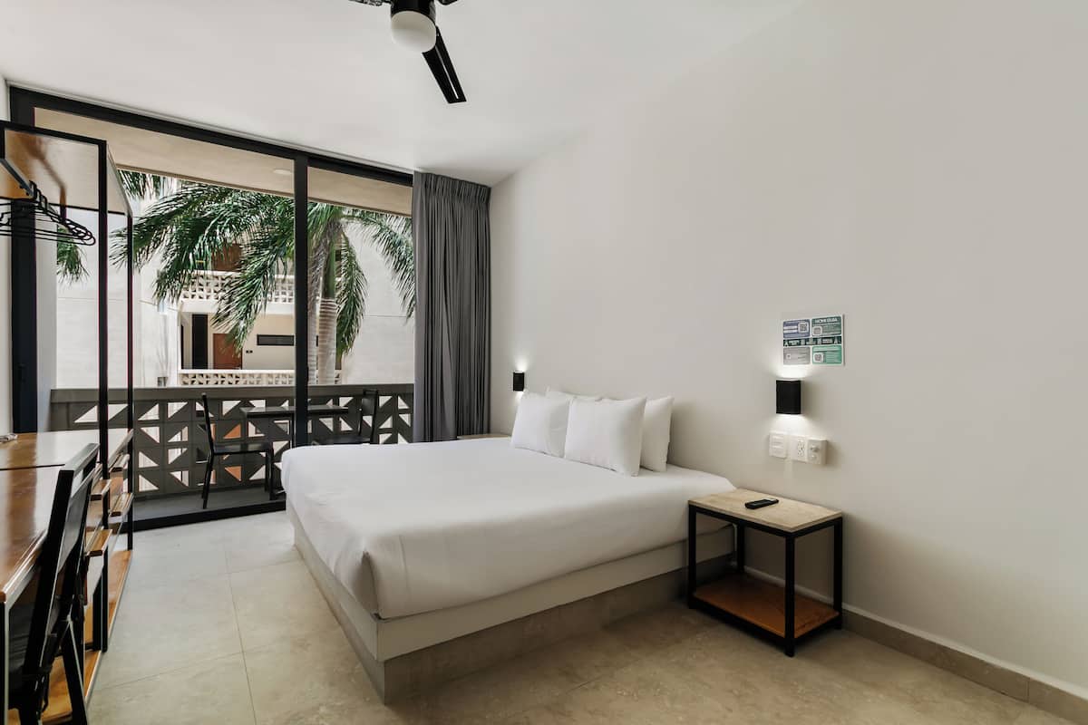 A hotel room with a bed, a desk, a chair, a small table, a nightstand, a wall-mounted light, and a view of palm trees and a building outside.