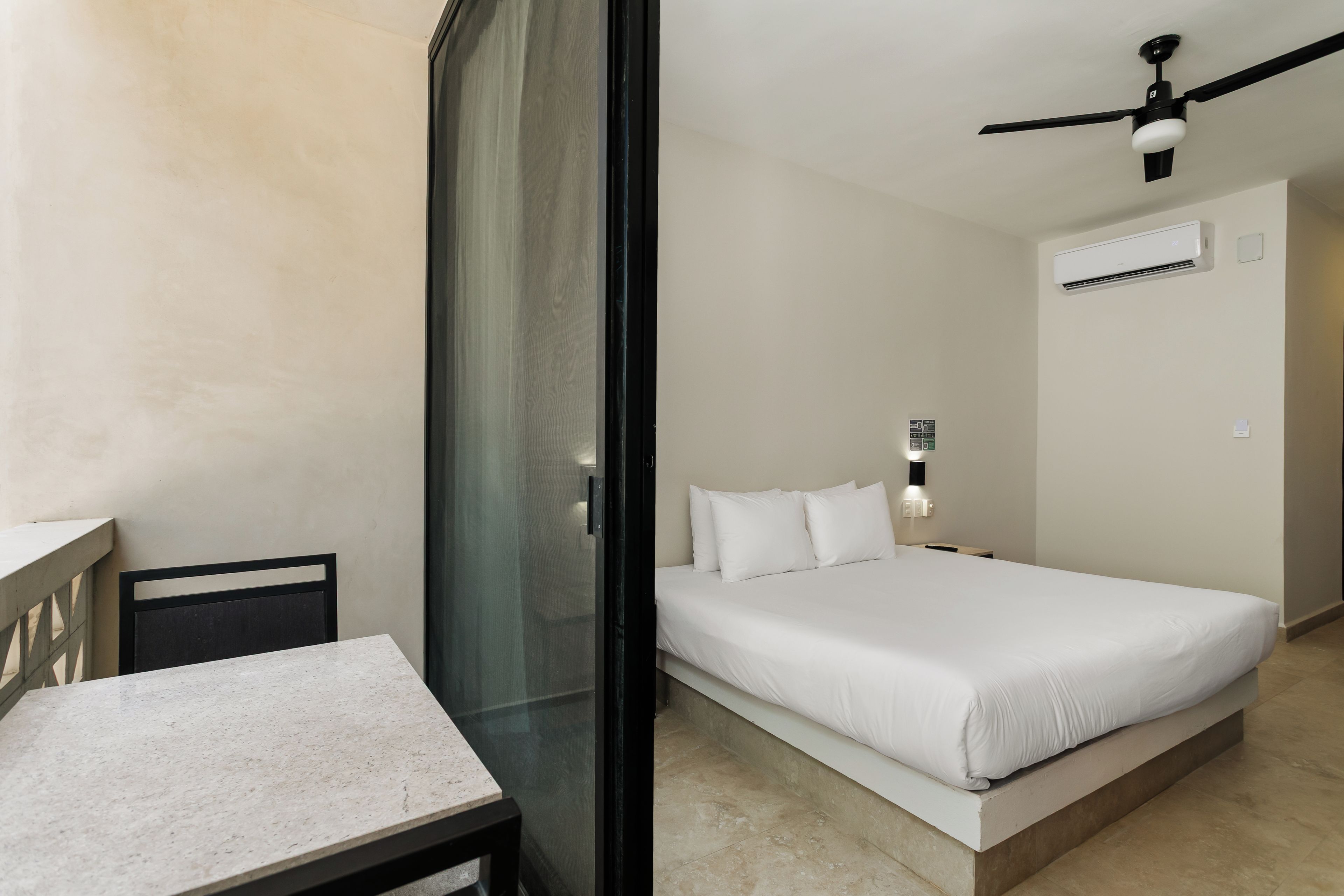 A bedroom with a bed, a table, a chair, a ceiling fan, and an air conditioning unit.