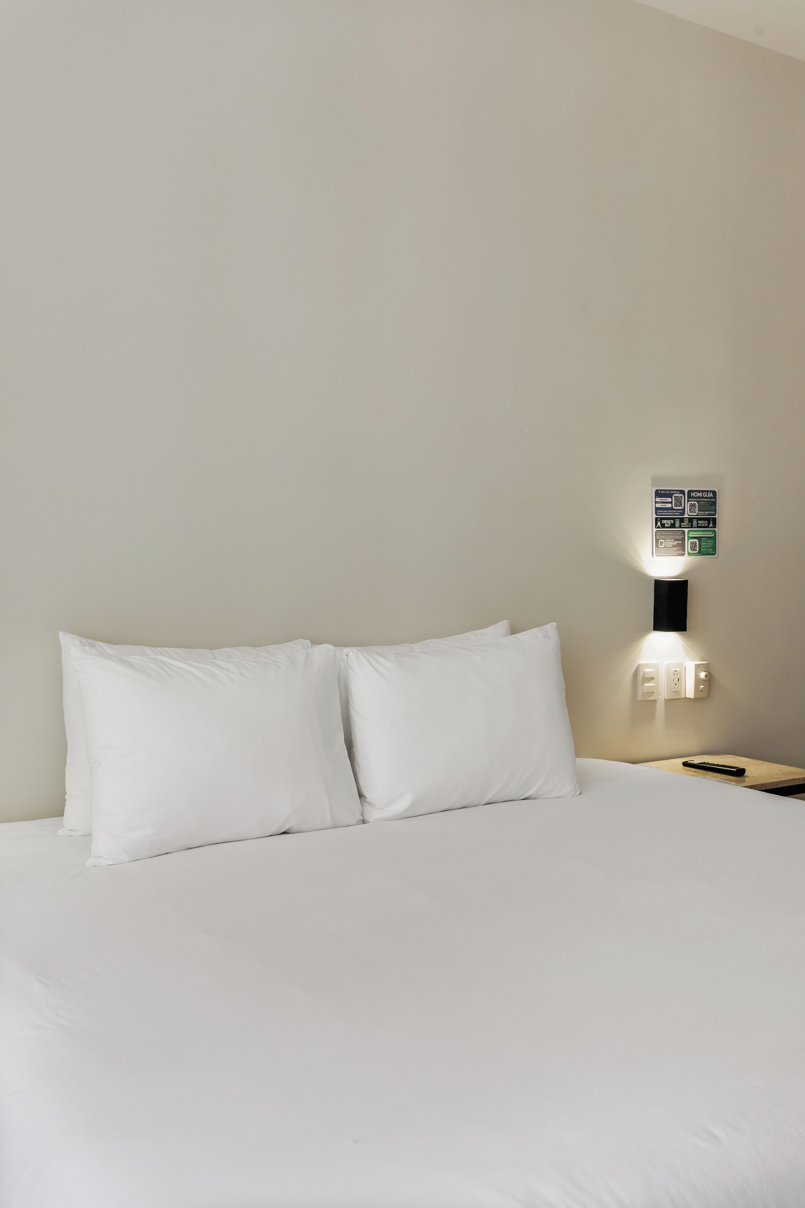 Une chambre d’hôtel avec un lit, des oreillers, une table de chevet, une télécommande et un luminaire fixé au mur.