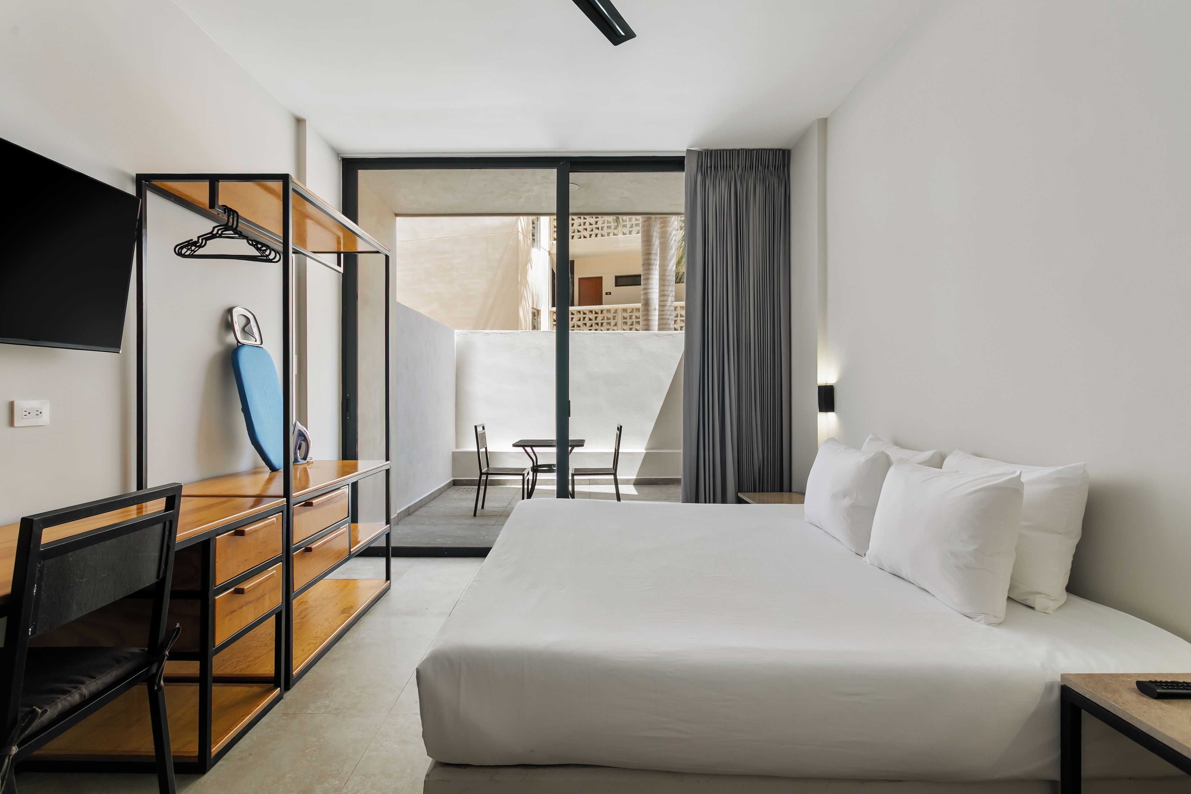 A hotel room with a bed, a desk with a chair, a TV mounted on the wall, and a balcony with a table and chairs.