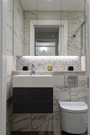 A bathroom with a marble wall, a sink with a modern faucet, a toilet, and a mirror above the sink. - Standard studio by Dmontrio (London)