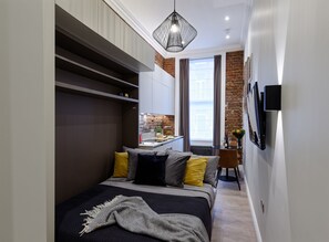 A narrow hotel room with a bed, a small kitchenette, a TV mounted on the wall, and a brick wall. - Standard studio by Dmontrio (London)