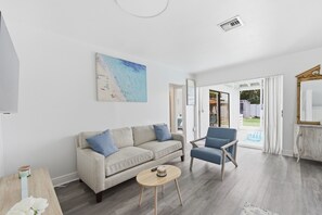 Ein Wohnzimmer mit einer Couch, einem Couchtisch und einem Sessel. An der Wand hängt ein Gemälde und ein Fenster mit Vorhängen.