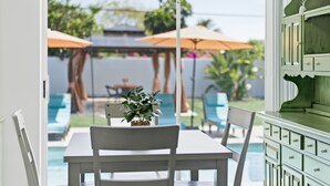 Comedor con una mesa, sillas, una planta en maceta y vistas a un área de piscina exterior con sombrillas.